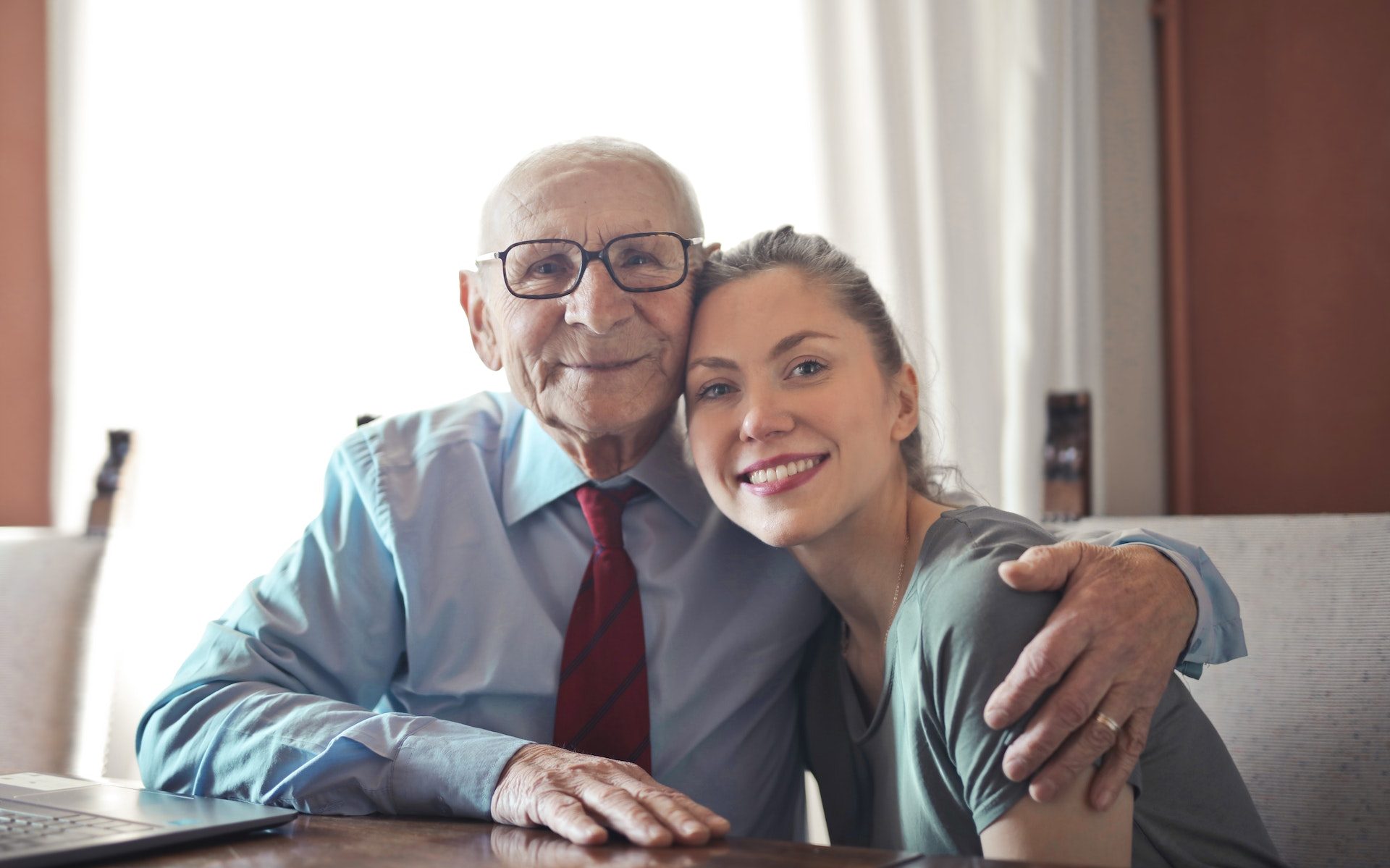 Positive senior man in formal wear and eyeglasses hugging with young lady while sitting at table